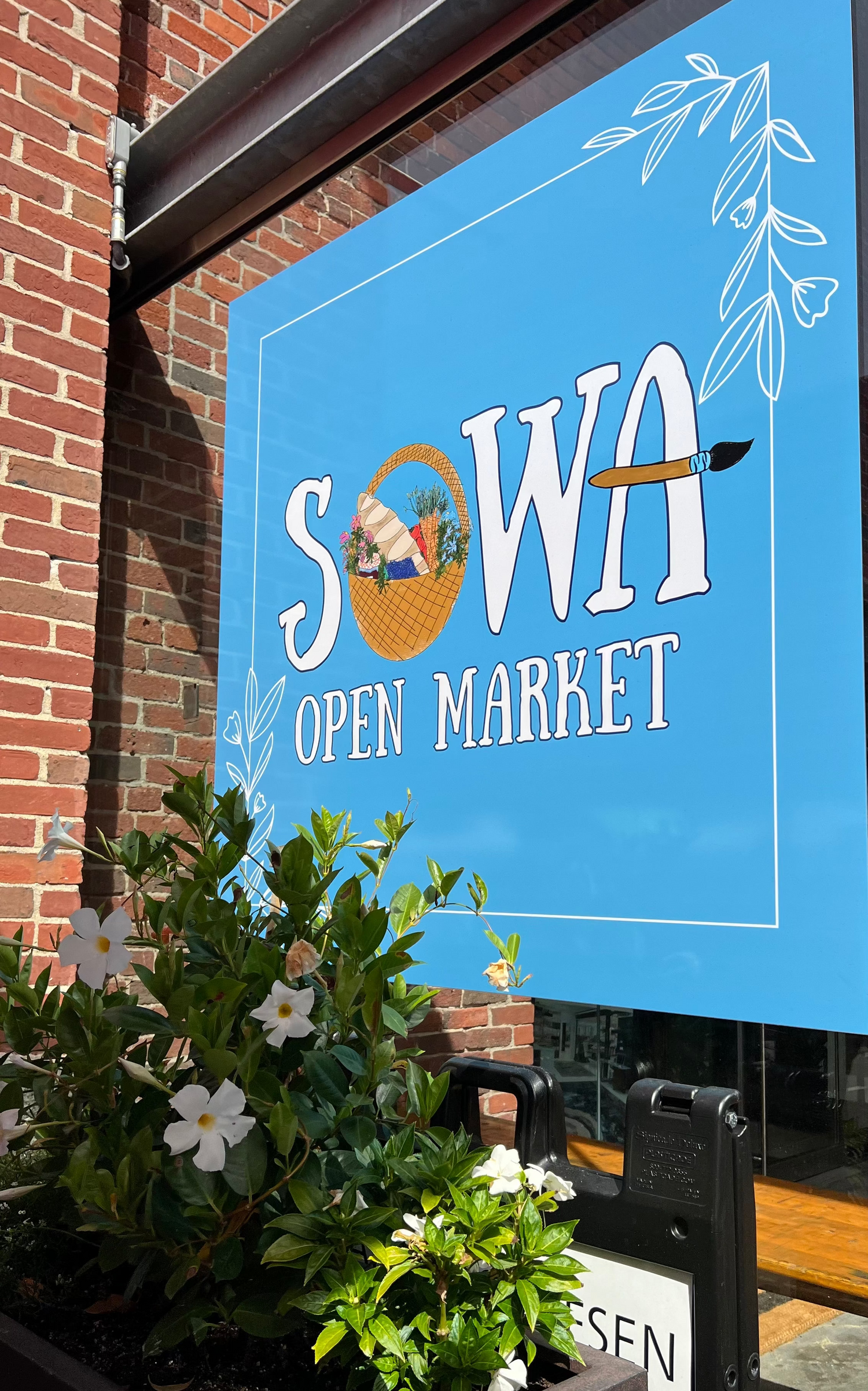A bright blue storefront sign reading 'SOWA OPEN MARKET' mounted on a brick building exterior, with white decorative leaf graphics and a planter box with green leaves and white flowers in the foreground.