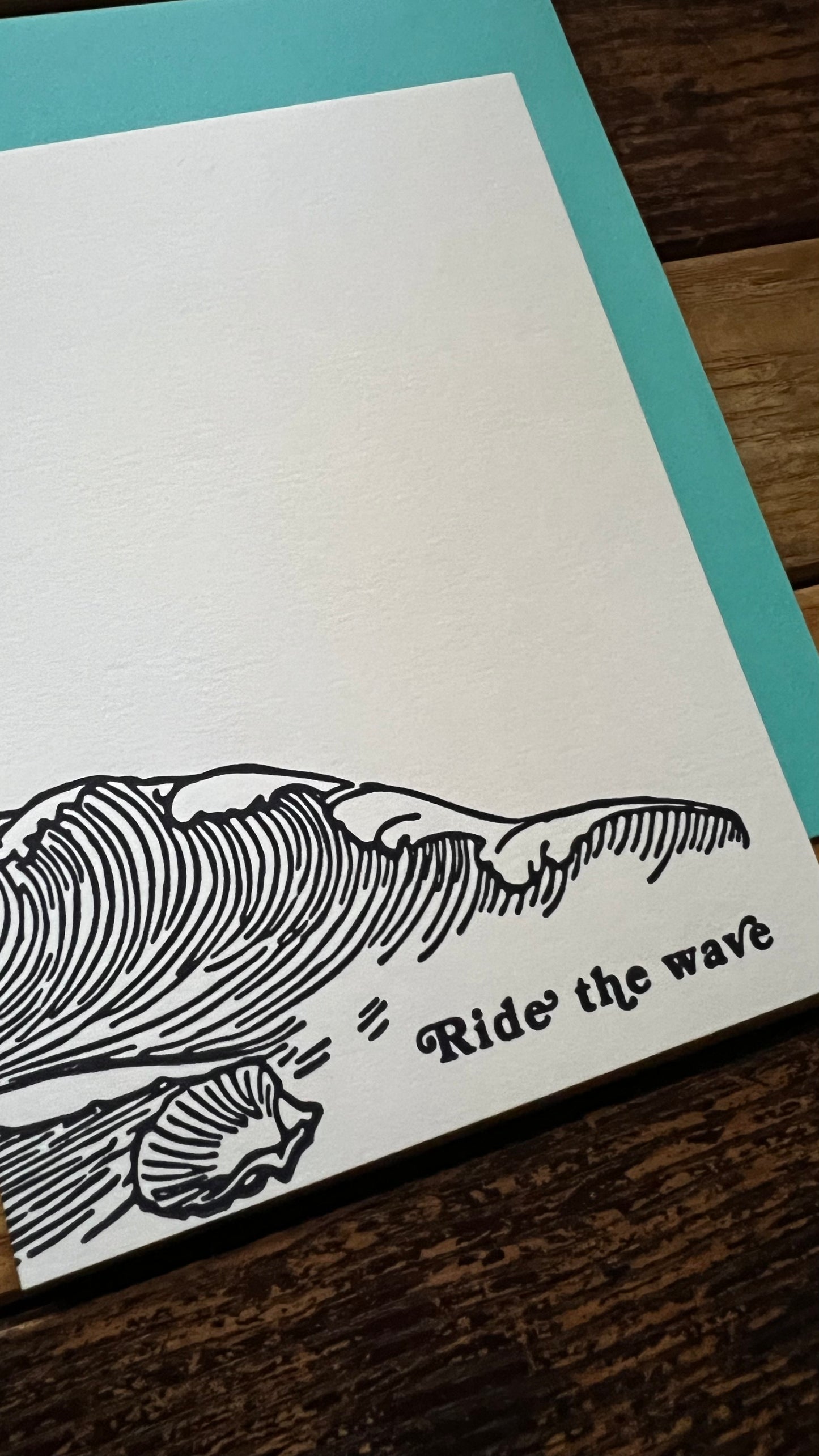 Note card with wave design and 'Ride the wave' text on a wooden surface.