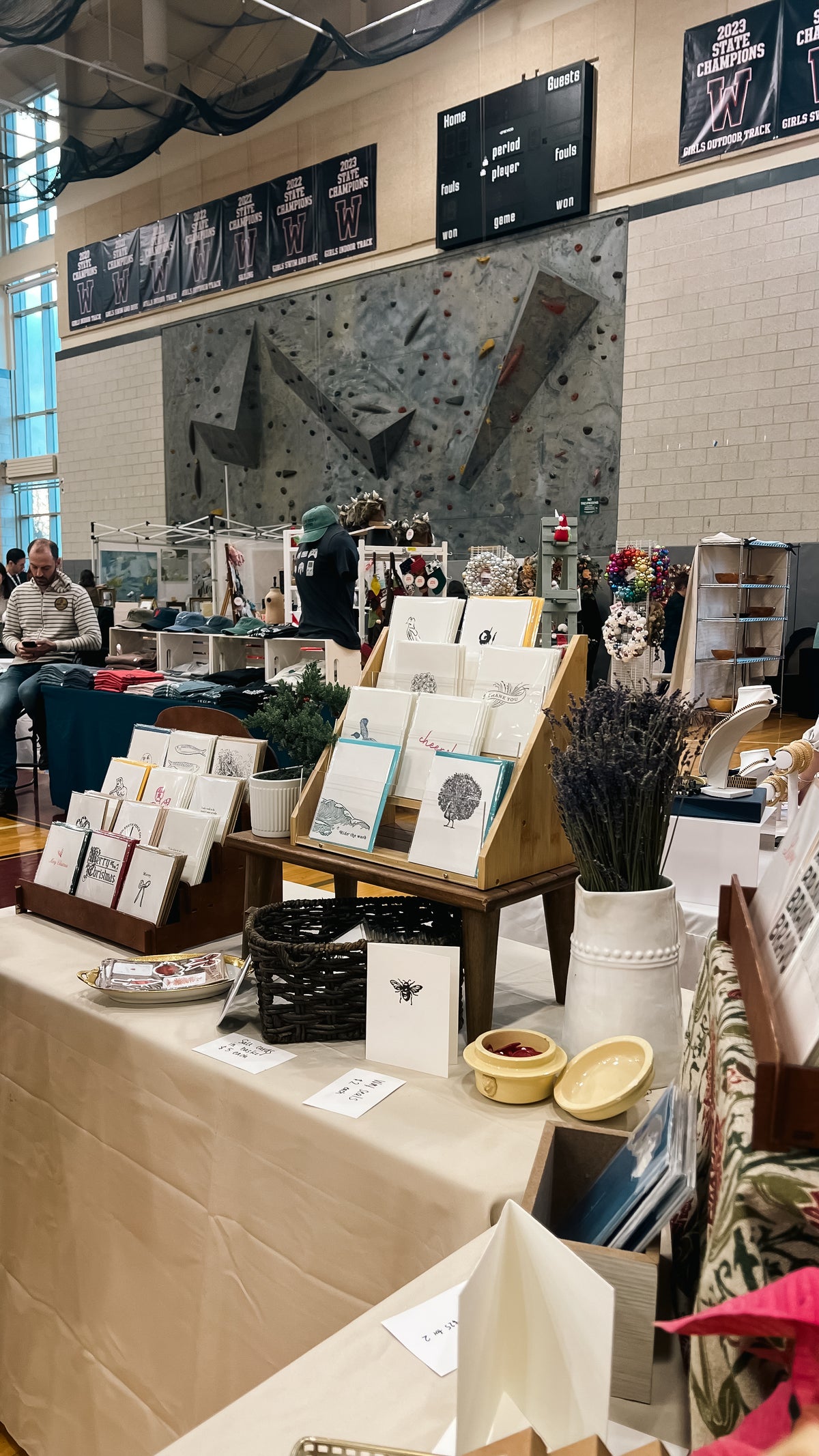 A craft fair booth displaying handmade greeting cards, small decorative items, and dried lavender in a beige tablecloth-covered table, with a climbing wall and indoor event space in the background.