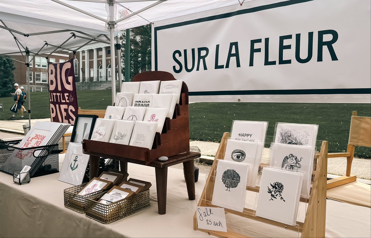 An outdoor market stall under a white canopy displaying handmade greeting cards, art prints, and signs. A large white banner in the background reads 'SUR LA FLEUR', and a smaller sign says 'BIG LITTLE PIES'.
