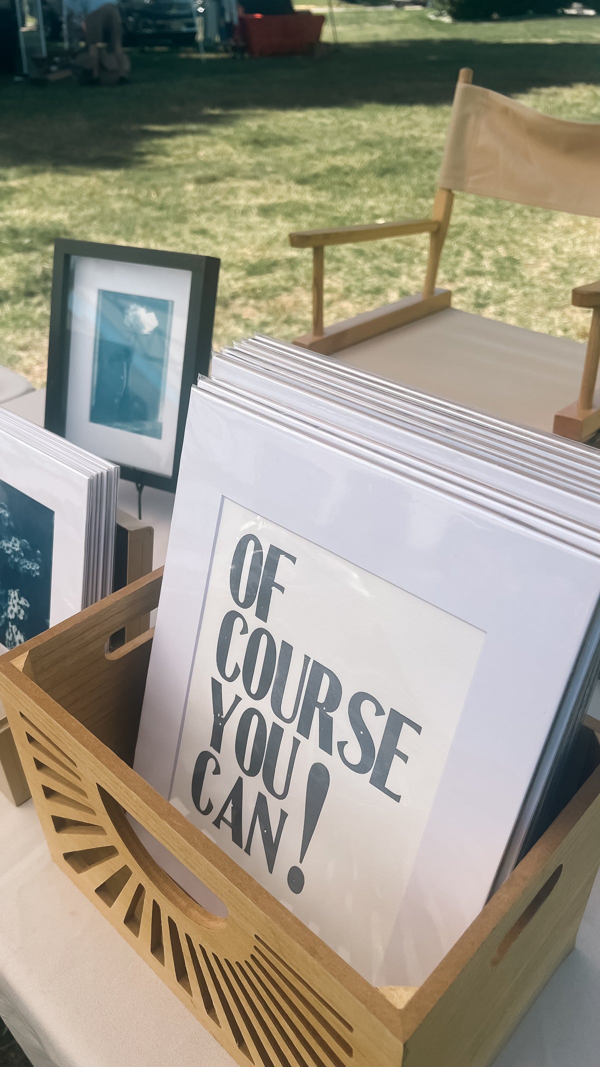 A wooden display tray holding a stack of framed art prints, with the front print reading 'OF COURSE YOU CAN!' in dark gray text on a white background. The scene is outdoors on grass with a chair and other framed pieces blurred in the background.
