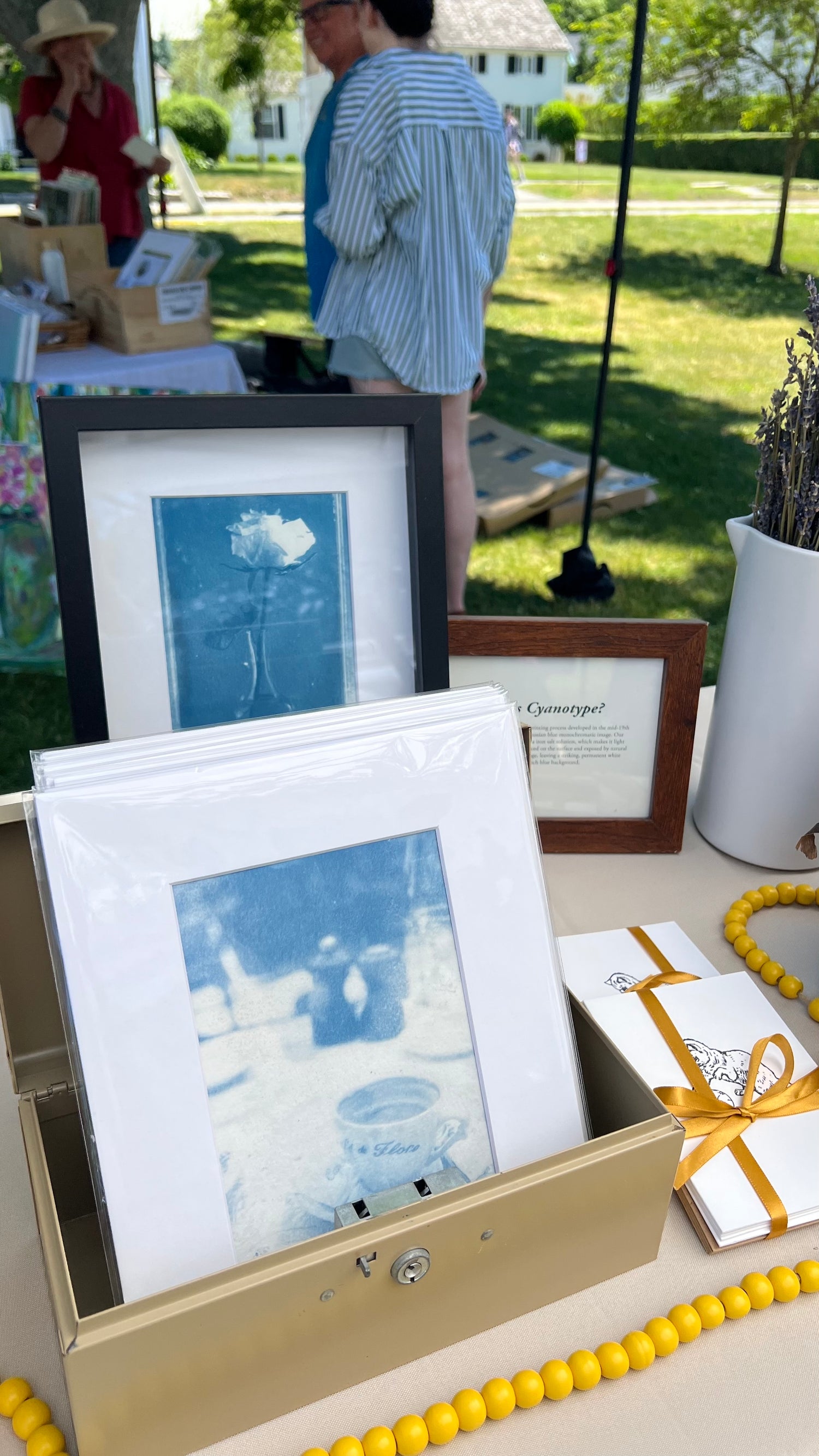 A display of blue-toned framed art prints and matted cards arranged on a table at an outdoor market. The products include a black-framed print, a blue-framed print, a matted card, and a small framed piece, with a gold box, yellow bead garland, and wrapped gift boxes nearby.